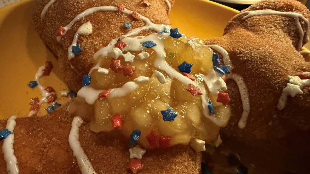 A close-up shot focusing on the topping of the patriotic Fourth of July Beignets. Diced, glossy apple pie filling sits nestled between the cinnamon-sugar-dusted pastries, heavily drizzled with white icing and garnished with small red, white, and blue star sprinkles.