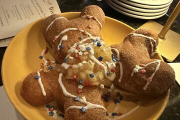 An angled, overhead view of three golden-brown, Mickey-shaped Fourth if July beignets served on a yellow plate. The treats are coated in cinnamon sugar, topped with a spoonful of warm apple compote, crisscrossed with white icing, and finished with red, white, and blue star sprinkles. A dim restaurant tabletop and an appetizer menu are visible in the background.