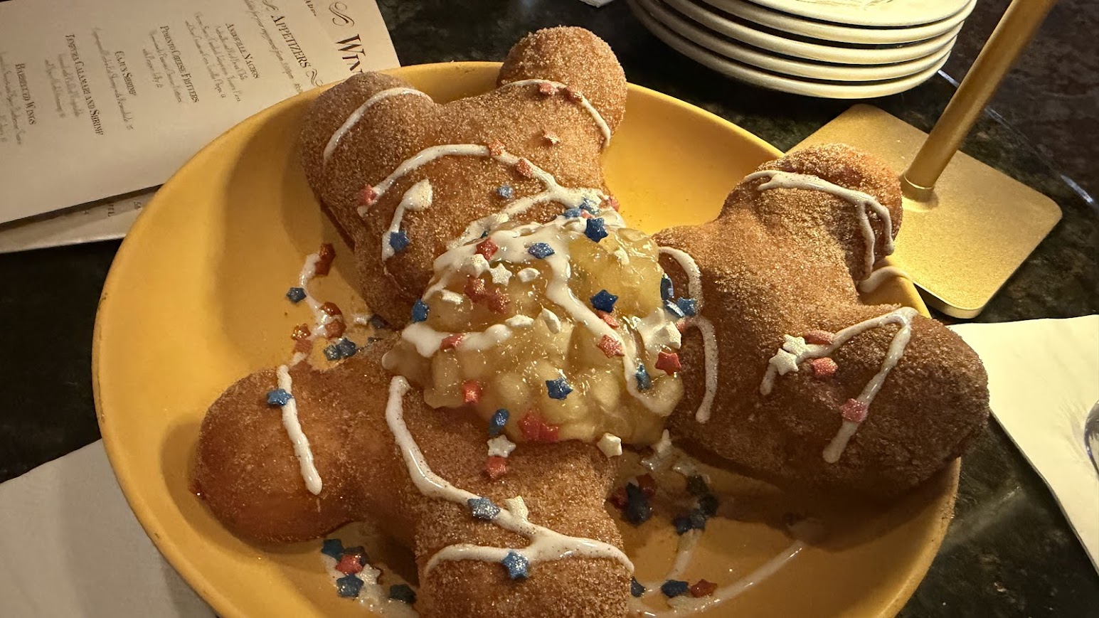 An angled, overhead view of three golden-brown, Mickey-shaped Fourth if July beignets served on a yellow plate. The treats are coated in cinnamon sugar, topped with a spoonful of warm apple compote, crisscrossed with white icing, and finished with red, white, and blue star sprinkles. A dim restaurant tabletop and an appetizer menu are visible in the background.