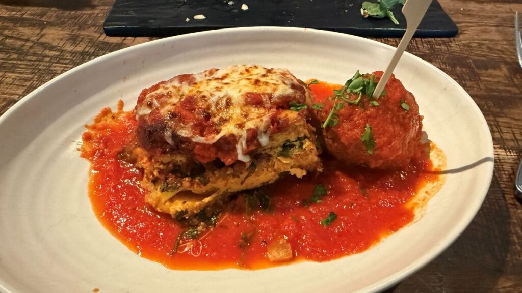 A thick slice of layered house-made lasagna topped with melted cheese and red sauce, served next to a large meatball with a toothpick on a pool of marinara sauce at Terralina Crafted Italian.