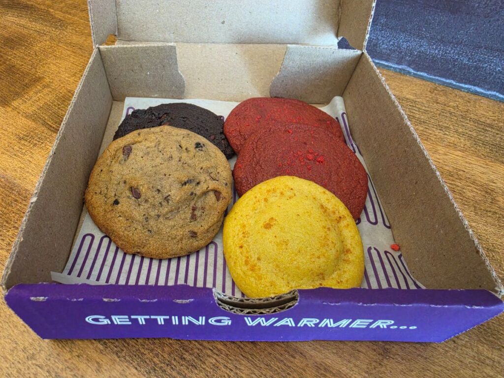 An open purple-and-white Insomnia Cookies cardboard box displaying five warm cookies on greaseproof paper, including a classic chocolate chip cookie, a double chocolate cookie, two vibrant red cookies, and one bright yellow cookie. The front flap reads "GETTING WARMER...".