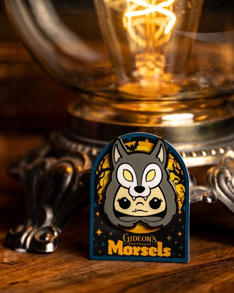 Colorful cartoon pin featuring a gray wolf over a cute human face, with 'Gideon's Bakehouse Morsels' text on a dark background.