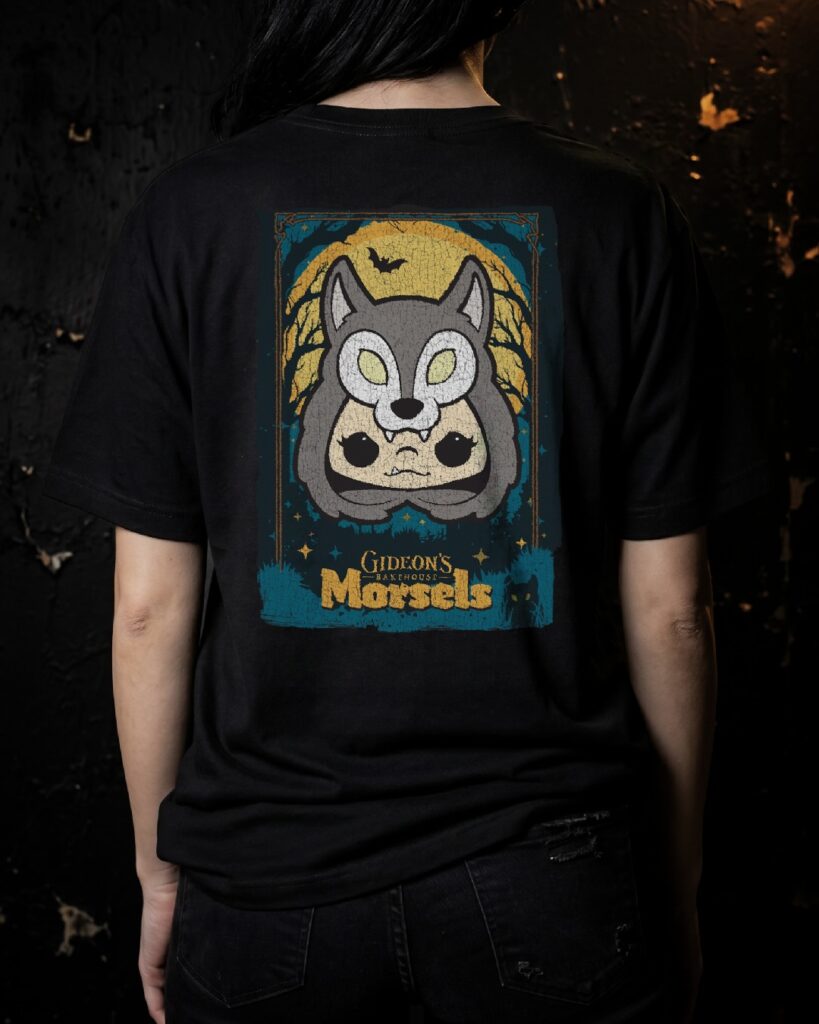 Back of a black T-shirt showing a framed, poster-style graphic: a gray wolf head over a yellow moon, with trees and the words 'Gideon's Bakehouse Morsels' at the bottom for Gideon's Bakehouse June 2026.