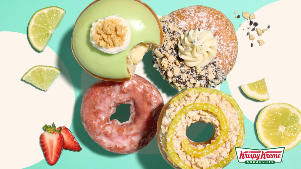 An overhead promotional shot of four seasonal Krispy Kreme summer doughnuts arranged against a bright turquoise and off-white abstract background. The top-left doughnut has pale green icing, a white cream star, and graham cracker crumbs on top, with a bite taken out, revealing a cream filling. To its right is a powdered doughnut topped with half chocolate sprinkles/crumbs and a swirl of buttercream frosting. The bottom-left doughnut is a classic ring with a translucent pink strawberry glaze, and the bottom-right doughnut is a ring topped with blonde biscuit crumbs and a bright yellow lemon drizzle spiral. Fresh lime slices, a cut lemon, and half a strawberry are scattered around the edges, with the Krispy Kreme logo visible in the bottom-right corner.