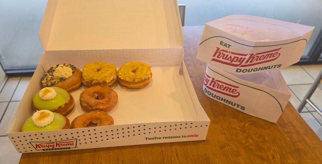 An open white Krispy Kreme box sitting on a wooden table filled with a colorful variety of Krispy Kreme Summer Doughnuts next to stacked paper bags.