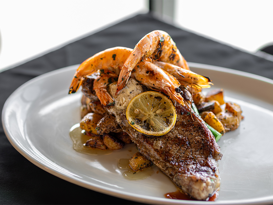 A close-up, high-angle shot of a surf and turf dish served on a white oval platter over a black tablecloth. A grilled New York strip steak is layered with roasted fingerling potatoes, topped with large, whole grilled head-on shrimp, a pat of herb butter, and a grilled lemon wheel.