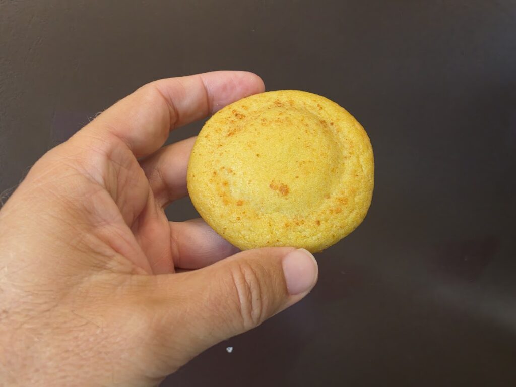 A hand holding a whole, round Lemon Solar Punch cookie, showing its smooth golden-yellow surface dusted with fine orange-yellow crumbles against a dark background.