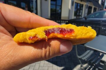 A close-up of a bright yellow Lemon Solar Punch cookie broken in half, revealing a thick, vibrant red fruit jam filling held in front of an outdoor patio background.