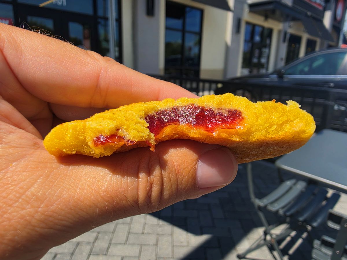 A close-up of a bright yellow Lemon Solar Punch cookie broken in half, revealing a thick, vibrant red fruit jam filling held in front of an outdoor patio background.
