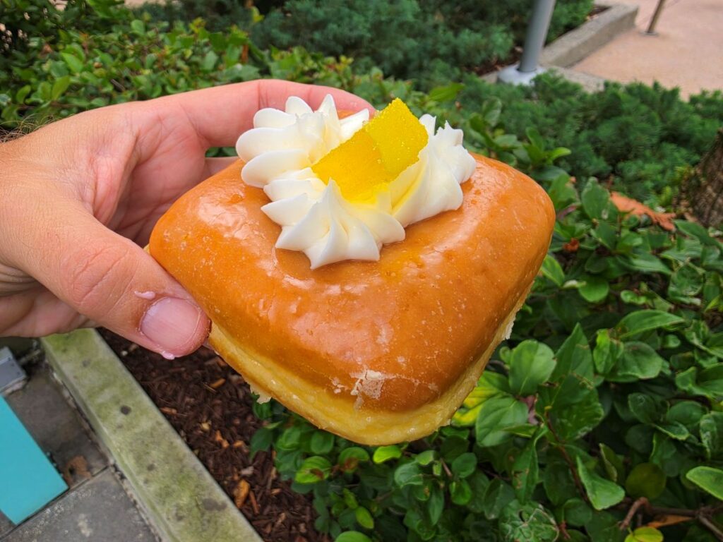 Hand holding a glossy glazed donut topped with white cream swirls and a small yellow candied zest, outdoors for Everglazed Disney Springs review