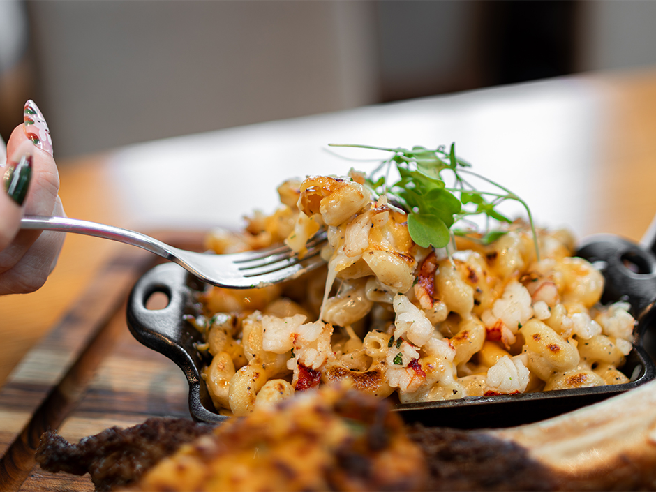 A detailed close-up shot of a hand with manicured nails lifting a forkful of cheesy lobster macaroni and cheese from a small, black cast-iron skillet. The dish is garnished with fresh microgreens, showcasing melted cheese pulls and visible chunks of lobster meat.