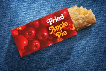 A McDonald's Fried Apple Pie resting on a blue denim background. The crispy, golden-brown fried crust has a bite taken out of the end, revealing the warm apple filling inside. The pie is partially tucked into a red sleeve adorned with an all-over bright red apple graphic and the words "Fried Apple Pie" written in a retro white and yellow script font.