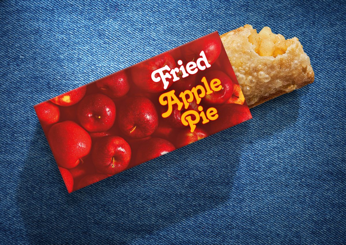 A McDonald's Fried Apple Pie resting on a blue denim background. The crispy, golden-brown fried crust has a bite taken out of the end, revealing the warm apple filling inside. The pie is partially tucked into a red sleeve adorned with an all-over bright red apple graphic and the words "Fried Apple Pie" written in a retro white and yellow script font.