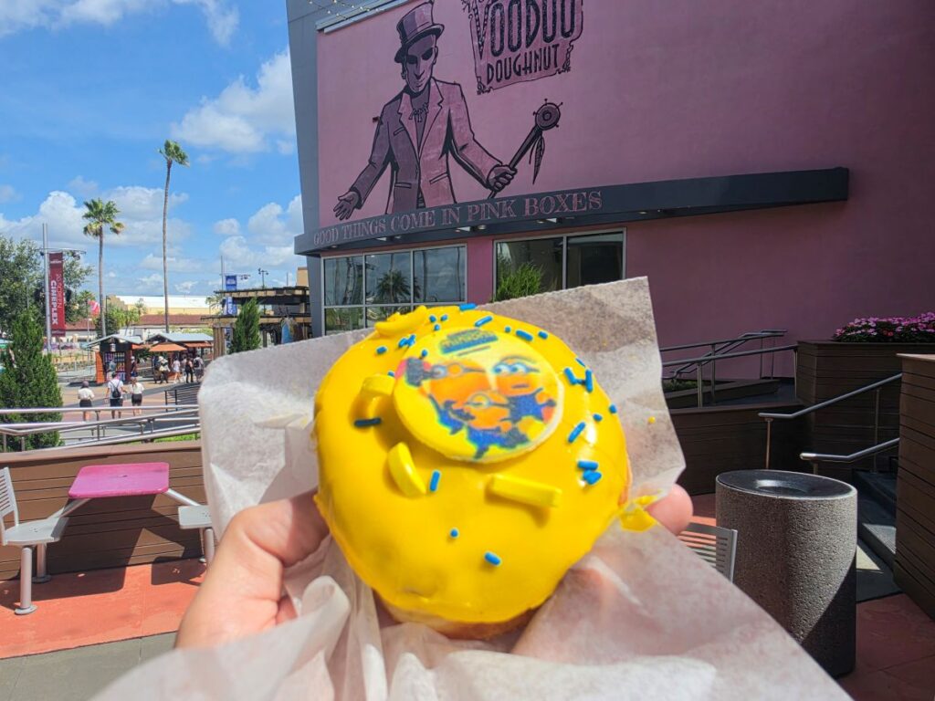 A person holding a colorful Minion Voodoo Doughnut in front of the pink Voodoo Doughnut shop at Universal CityWalk on a bright, clear day.