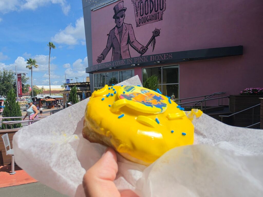 A side angle of the Minion Voodoo Doughnut held outside the pink Voodoo Doughnut storefront, showing the thick profile of the yeast doughnut and its glossy yellow icing.