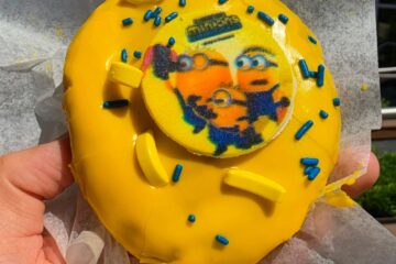 A detailed top-down view of the Minion Voodoo Doughnut being held in sunlight, highlighting the texture of the edible image disc, scattered blue sprinkles, and small yellow banana candy toppings on the thick yellow icing.