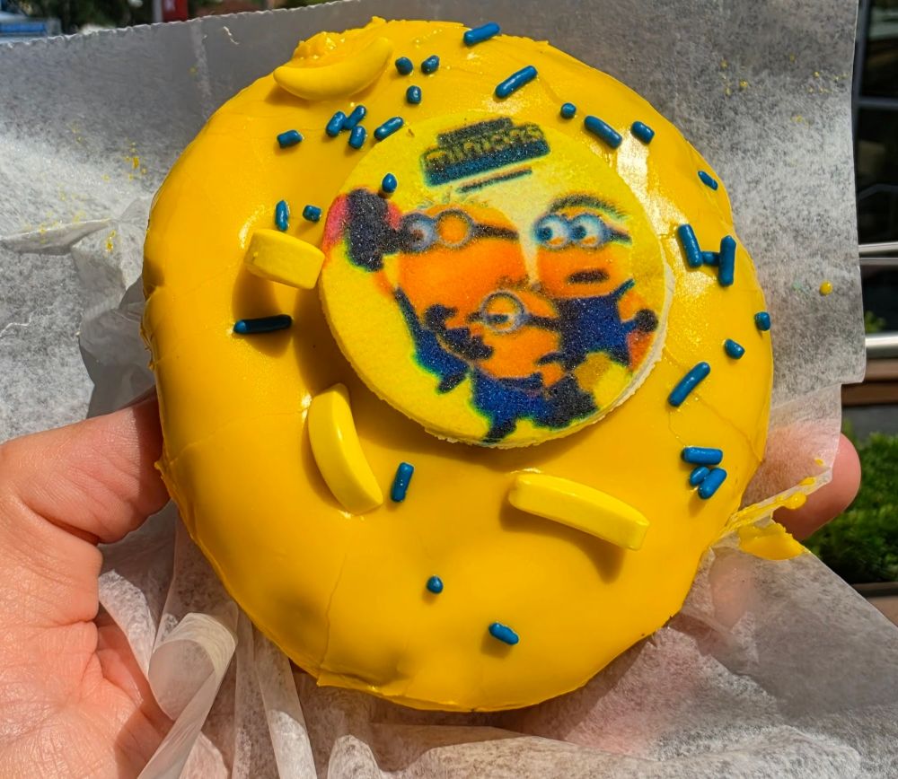 A detailed top-down view of the Minion Voodoo Doughnut being held in sunlight, highlighting the texture of the edible image disc, scattered blue sprinkles, and small yellow banana candy toppings on the thick yellow icing.