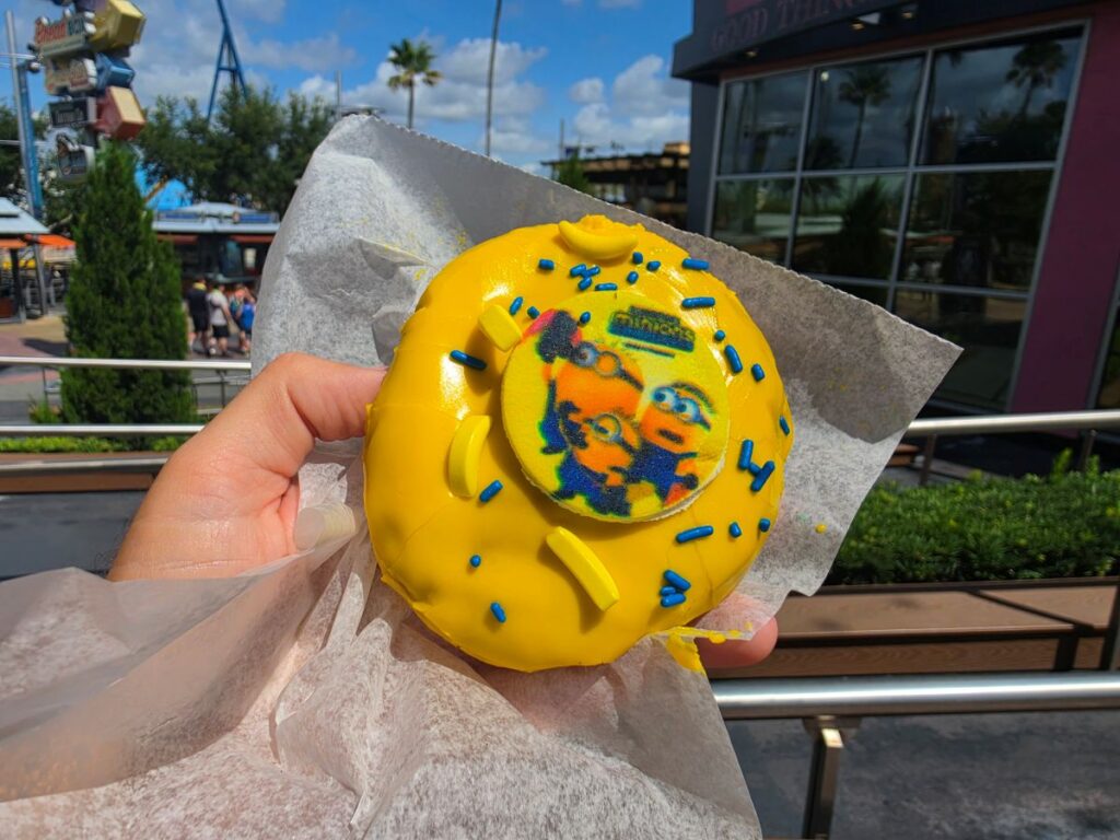 A hand holds up a Minions Voodoo Doughnut at Universal CityWalk, with a theme park coaster track and palm trees visible under a blue sky with white clouds in the background.