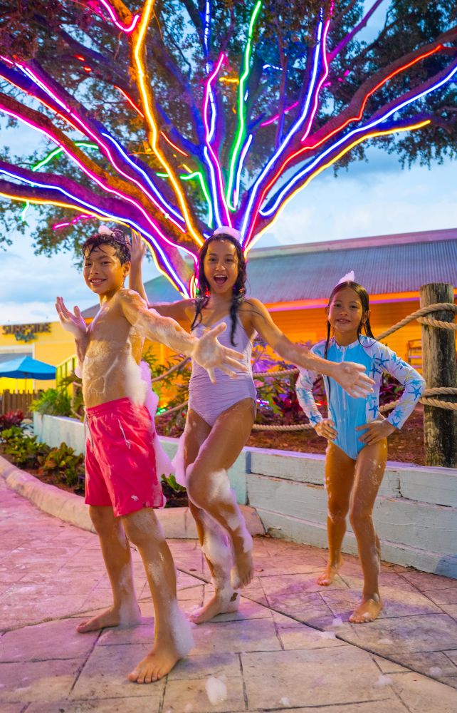 Three children covered in party foam dancing outside on a patio beneath a large tree decorated with bright, glowing neon lights at AquaGlow Adventure Island Tampa.