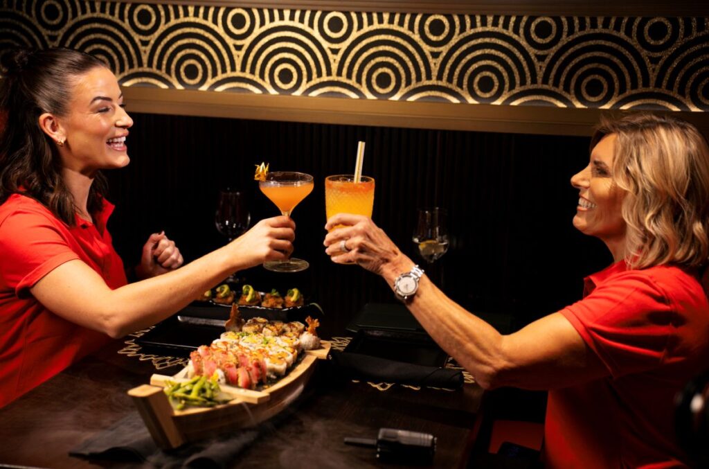 Two women in red shirts smiling and raising their cocktail glasses in a toast over a table featuring a P.F. Chang's Sushi Boat surrounded by ambient, swirling smoke.