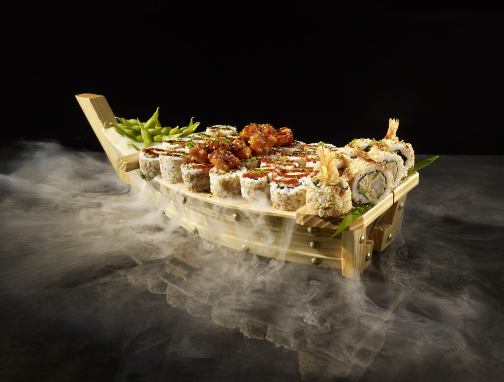 A dramatic studio shot of a P.F. Chang's Sushi Boat laden with an assortment of sushi rolls, crispy shrimp tempura rolls, and a side of edamame, completely enveloped in dense white smoke over a reflective black surface.
