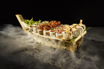 A dramatic studio shot of a P.F. Chang's Sushi Boat laden with an assortment of sushi rolls, crispy shrimp tempura rolls, and a side of edamame, completely enveloped in dense white smoke over a reflective black surface.