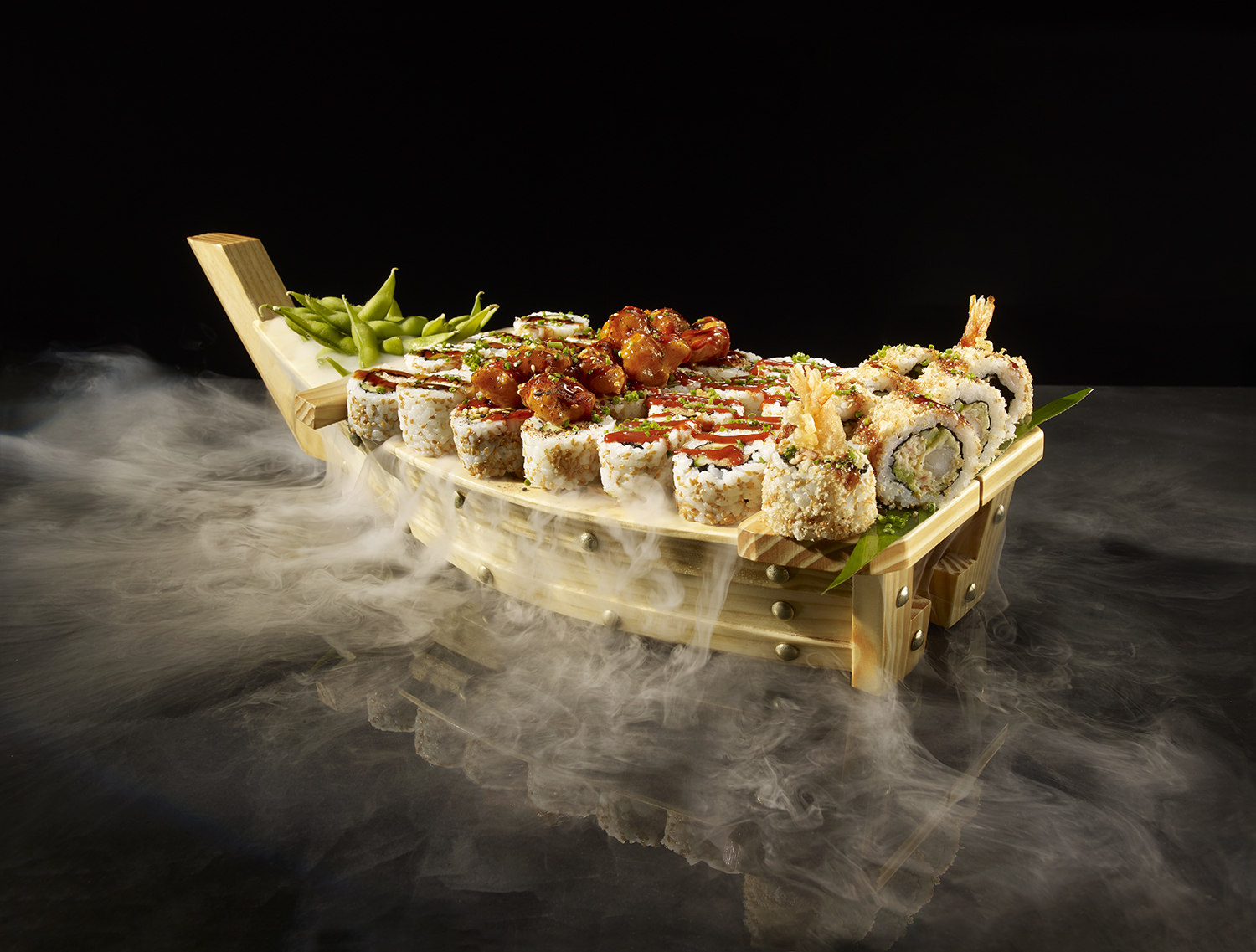 A dramatic studio shot of a P.F. Chang's Sushi Boat laden with an assortment of sushi rolls, crispy shrimp tempura rolls, and a side of edamame, completely enveloped in dense white smoke over a reflective black surface.