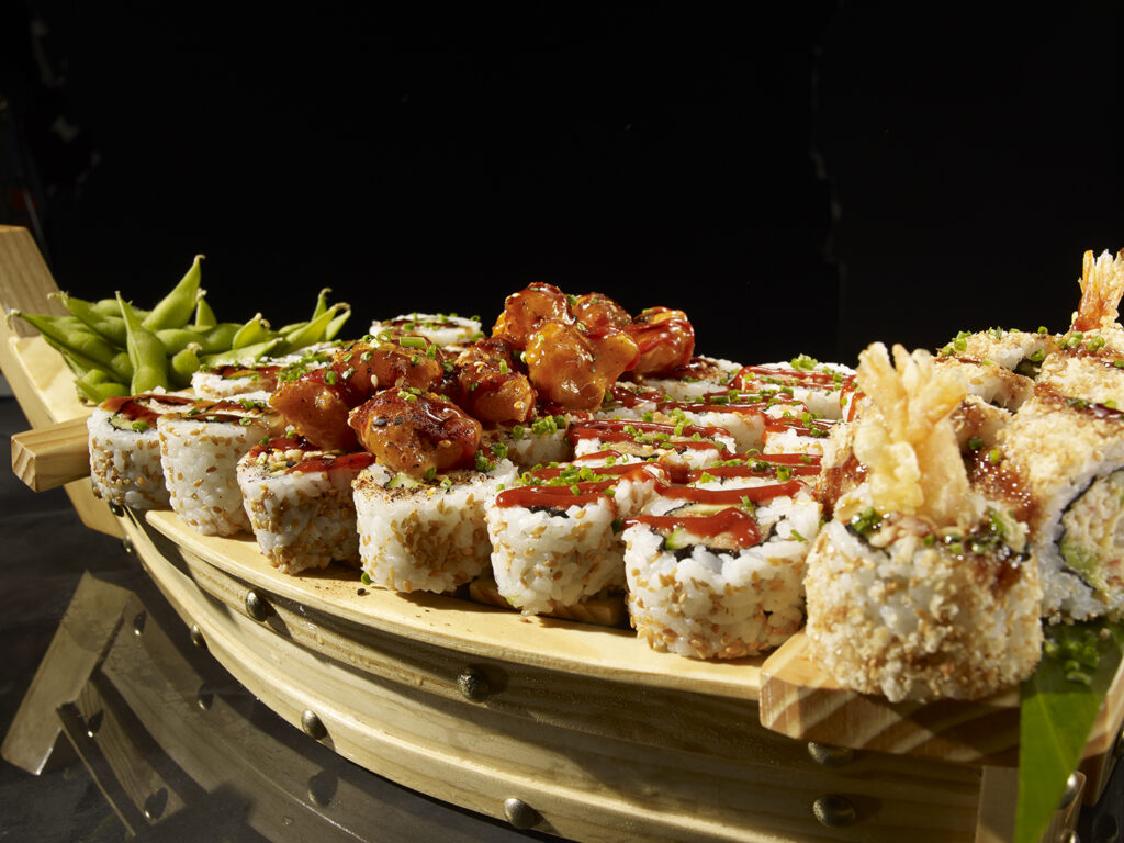 A close-up, detailed shot of a P.F. Chang's Sushi Boat highlighting rows of variety sushi rolls drizzled with savory sauces, crispy shrimp tempura tails, and a mound of green edamame pods at the bow.