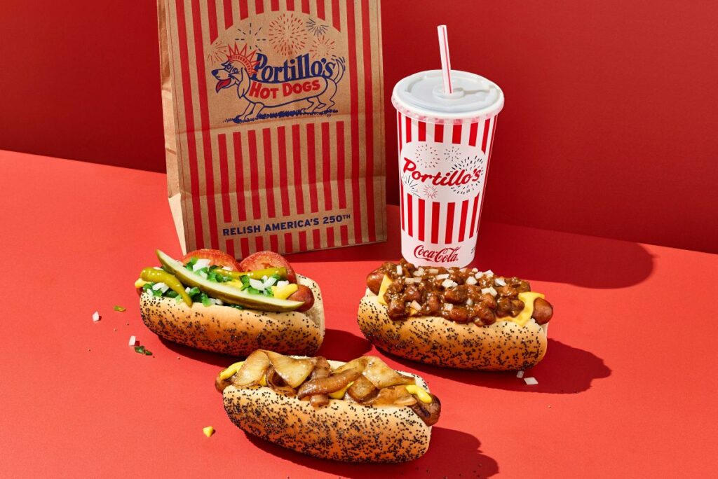 A Portillo's meal spread on a solid red background. It features a classic Chicago-style hot dog with pickles and tomatoes, a chili cheese dog with diced onions, and a hot dog topped with grilled onions and mustard, all on poppy seed buns. A red-and-white striped paper bag reading "RELISH AMERICA'S 250TH" and a matching striped Coca-Cola cup sit in the background.