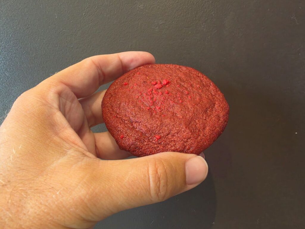 A close-up shot of a hand holding a Supergirl Insomnia Cookies whole, vibrant red Red Velvet Revenge cookie outdoors, showing a rich texture lightly sprinkled with red velvet cake crumbs.