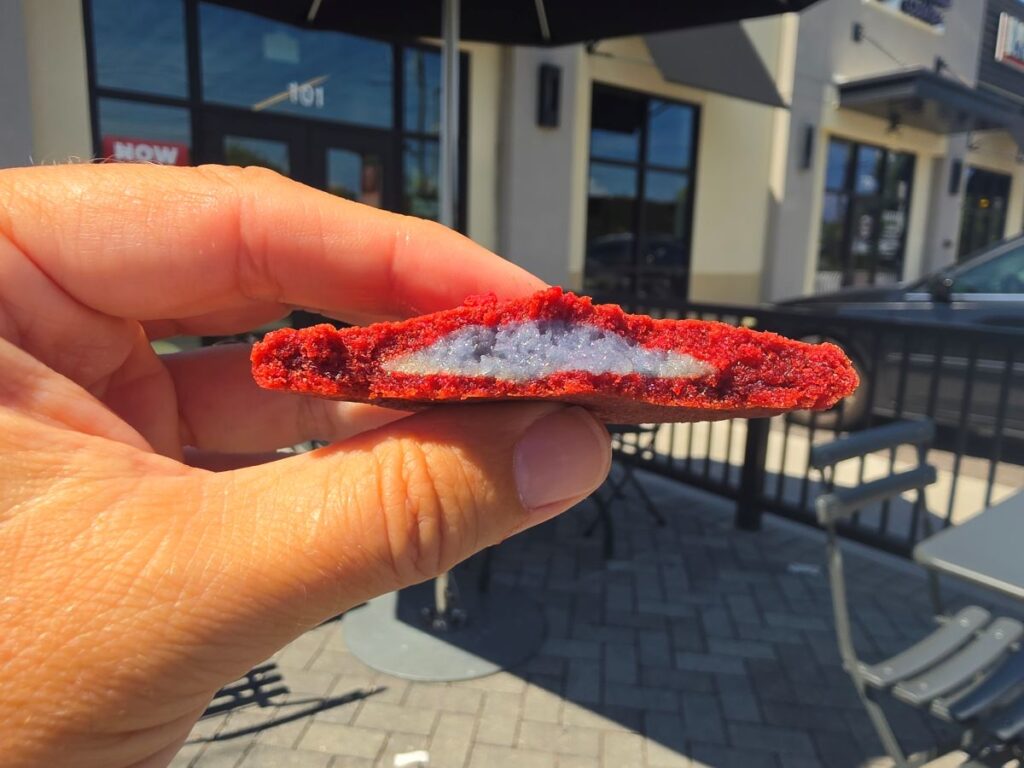A close-up of a vibrant red Red Velvet Revenge cookie split in half, showcasing a thick, contrasting light blue-grey frosting filling inside for the Supergirl Insomnia Cookies