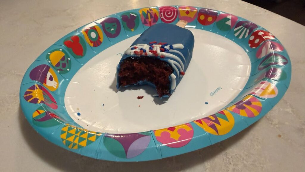 A wide shot of a blue, popsicle-shaped Fourth of July cake pop with a large bite missing from the front. The treat sits on a light blue Disney paper plate decorated with stylized theme park icons, placed on a light-colored countertop for a Disney cake pop review