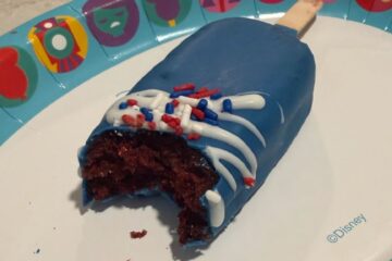 A close-up, high-angle shot of a popsicle-shaped cake pop with a bite taken out of it, revealing a moist red velvet interior. The treat is coated in vibrant blue icing with a white drizzle and red, white, and blue sprinkles, resting on a Disney-themed paper plate.