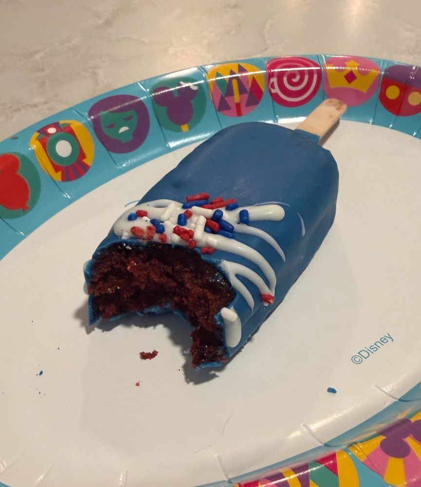 A close-up, high-angle shot of a popsicle-shaped cake pop with a bite taken out of it, revealing a moist red velvet interior. The treat is coated in vibrant blue icing with a white drizzle and red, white, and blue sprinkles, resting on a Disney-themed paper plate.