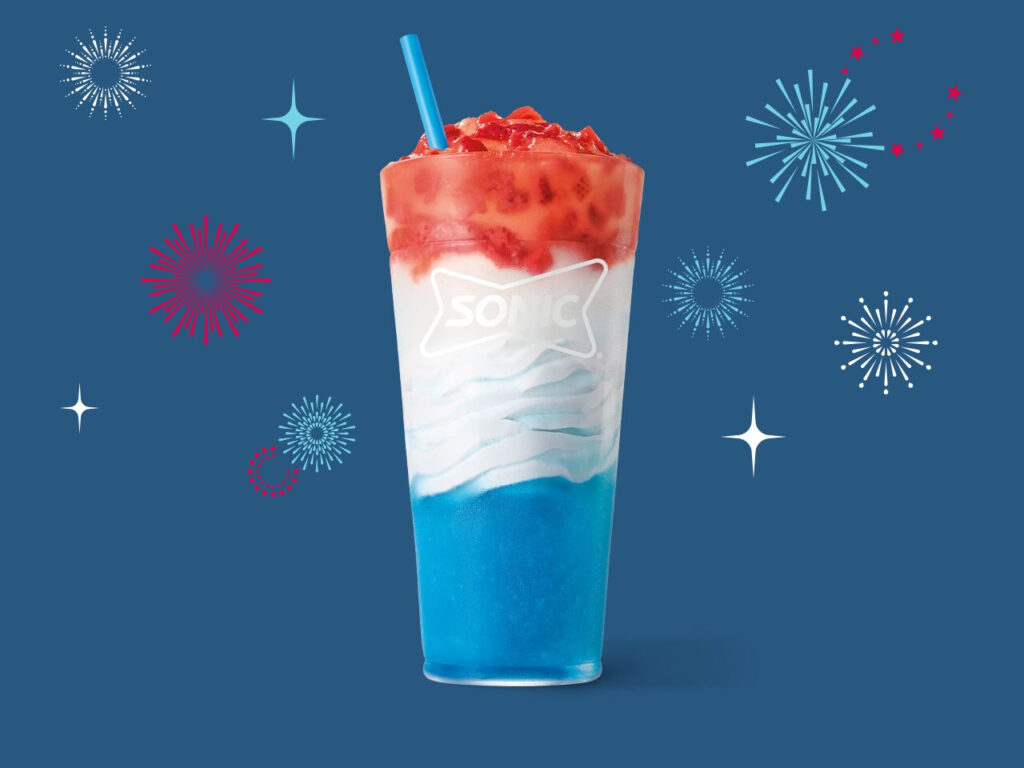 A tall, clear cup showcasing Sonic's Red, White & Blue Slush Float against a dark blue background decorated with red, white, and blue firework and star graphics. The frozen treat features distinct patriotic layers: a vibrant blue slush at the bottom, a white vanilla soft serve middle layer with a white Sonic logo on the cup, and a bright red strawberry topping with a blue straw sticking out.