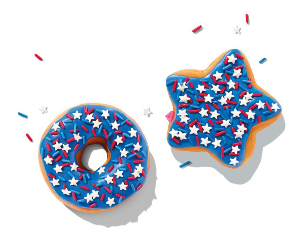 Two festive Dunkin Fourth of July donuts resting on a clean white background. On the left is a classic ring donut with bright blue icing, covered in red sprinkles and tiny white star sprinkles. On the right is a star-shaped filled donut featuring the same blue icing, red sprinkles, and white star sprinkle pattern.