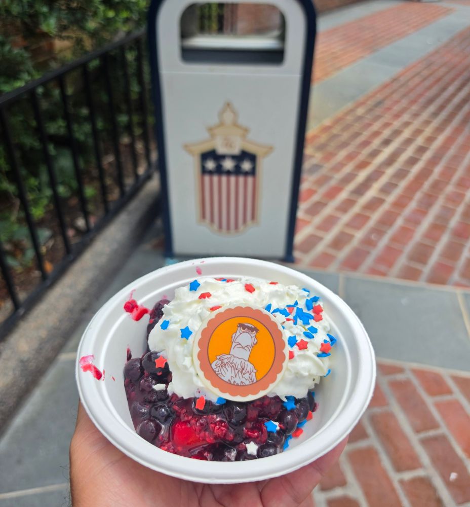 Close-up of Fourth of July Treats featuring a star-spangled berry shortcake dessert with a Sam Eagle chocolate disc, held in a hand against a backdrop of green garden bushes.