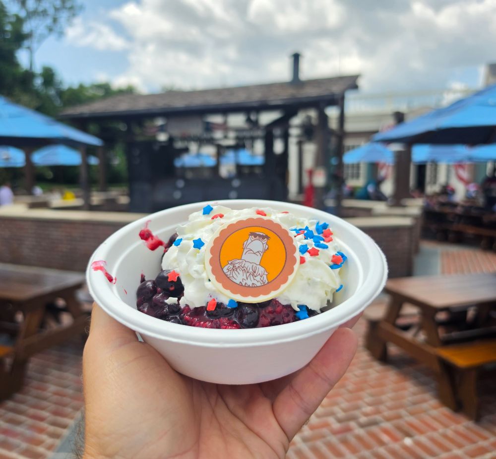 An angled shot of a hand holding a patriotic berry shortcake dessert with whipped cream and star sprinkles, highlighting delicious Fourth of July Treats at an outdoor dining location.