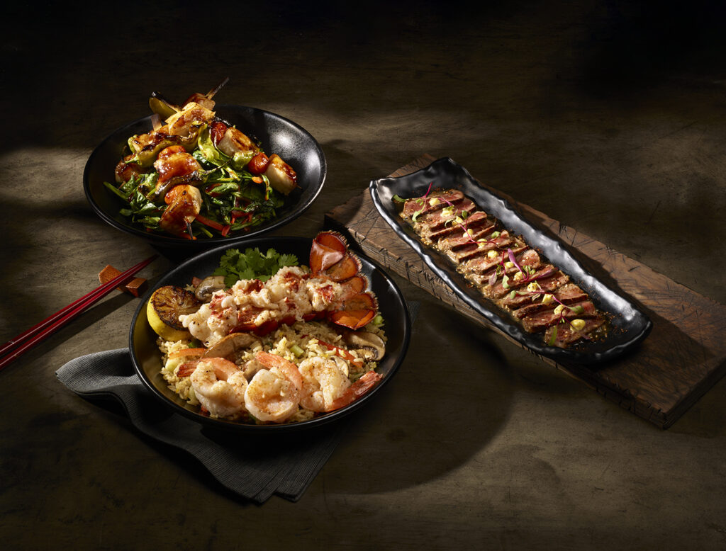 An elegant spread of three dishes featuring a bowl of fried rice topped with shrimp and lobster tail, a bowl of skewered sea bass over greens, and a long plate of sliced steak drizzled with sauce.