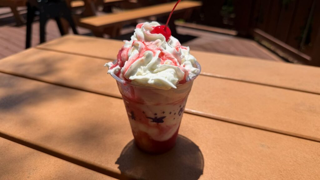 A clear plastic cup containing a Strawberry Shortcake dessert layered with cake, strawberry puree, swirl ice cream, whipped cream, and a cherry on top - one of the two Blizzard Beach treats