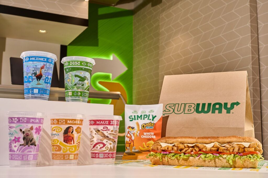 A Subway Moana Meal Deal promotional display of a Subway meal deal featuring five Disney Moana collectible plastic cups stacked on the left. On the right, a footlong grilled chicken submarine sandwich sits next to a bag of Simply Cheetos Puffs White Cheddar and a classic brown Subway paper bag.
