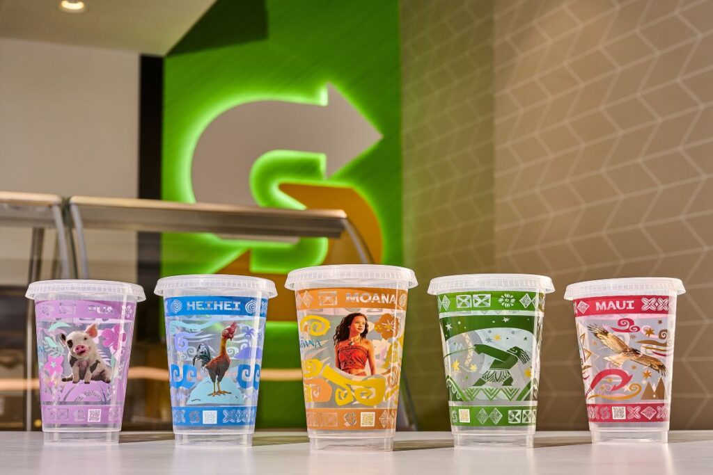A row of five clear Subway plastic collectible cups featuring colorful graphic designs of characters from Disney’s Moana. From left to right, the cups showcase Pua the pig on a purple background, Heihei the rooster on a blue background, Moana on an orange background, Maui in his human form on a green background, and Maui in his hawk form on a red background. The bright green Subway logo is visible in the blurred background.