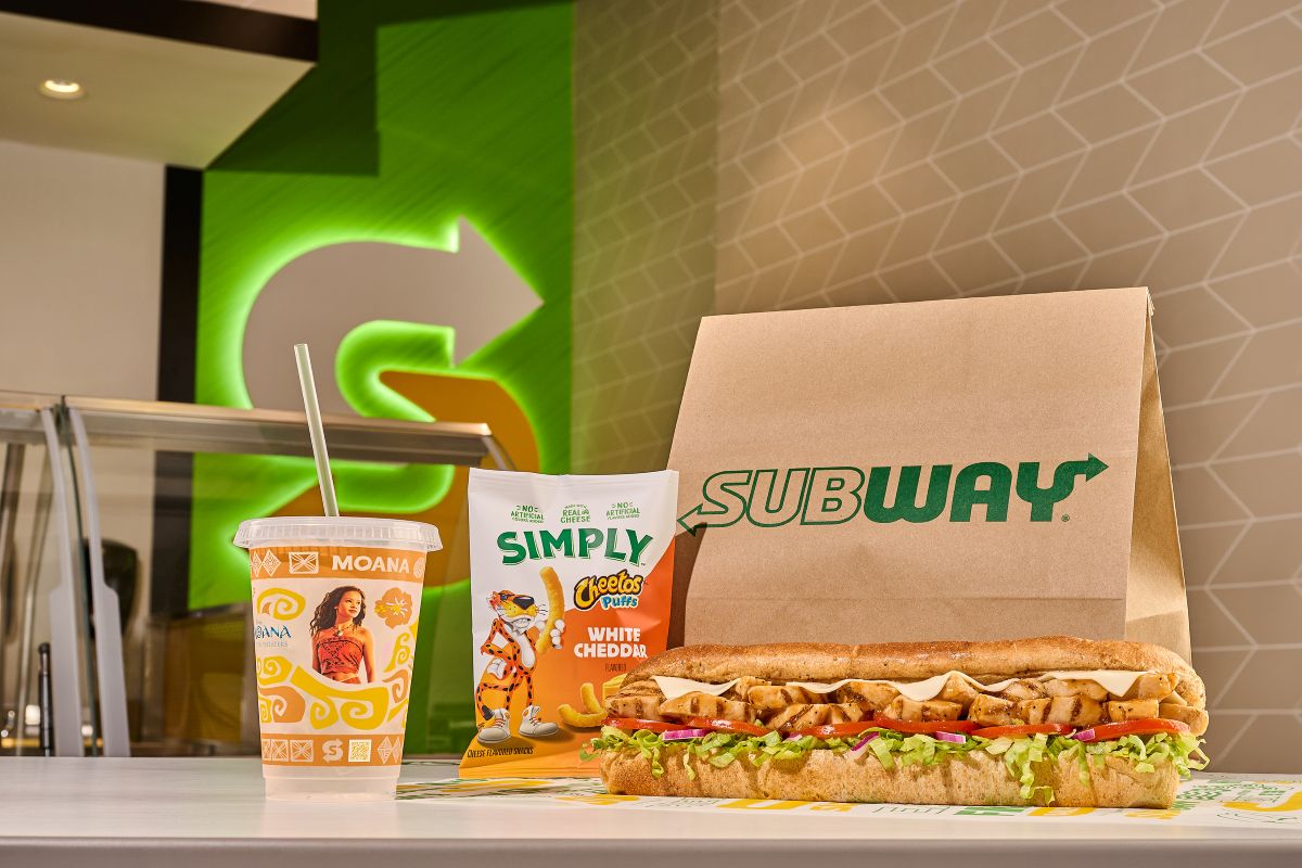 A Subway Moana meal deal layout featuring a footlong grilled chicken sub with lettuce, tomatoes, red onions, and melted cheese on a counter. Next to the sandwich is a bag of Simply Cheetos Puffs White Cheddar, a brown Subway paper bag, and a clear Moana collectible cup with a green straw, all set against a blurred green Subway logo background.