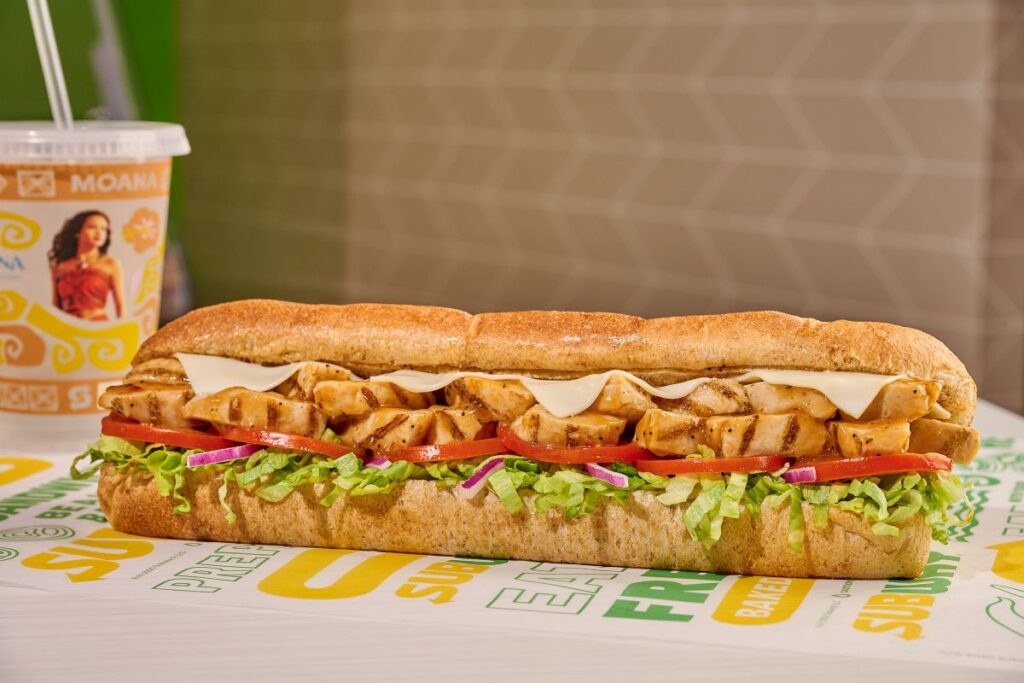A close-up shot of a Subway footlong grilled chicken sandwich on branded deli paper, layered with melted white cheese, sliced tomatoes, shredded lettuce, and red onions. A clear plastic Disney Moana collectible cup with a white straw is partially visible in the soft-focus background.