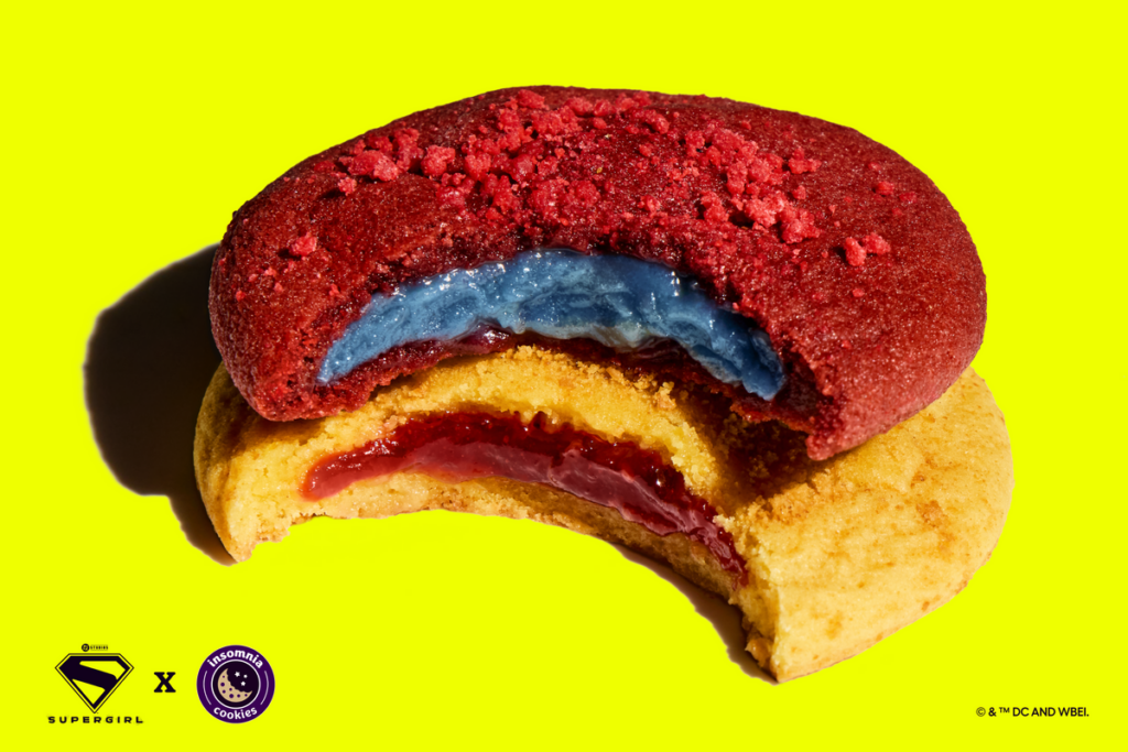 A top-down close-up shot of two halved cookies stacked on top of each other against a solid bright yellow background. The top cookie is a Red Velvet Revenge cookie with a blue dough center, and the bottom is a Lemon Solar Punch cookie with a red jam center. Small Supergirl and Insomnia Cookies logos are visible in the bottom left corner.