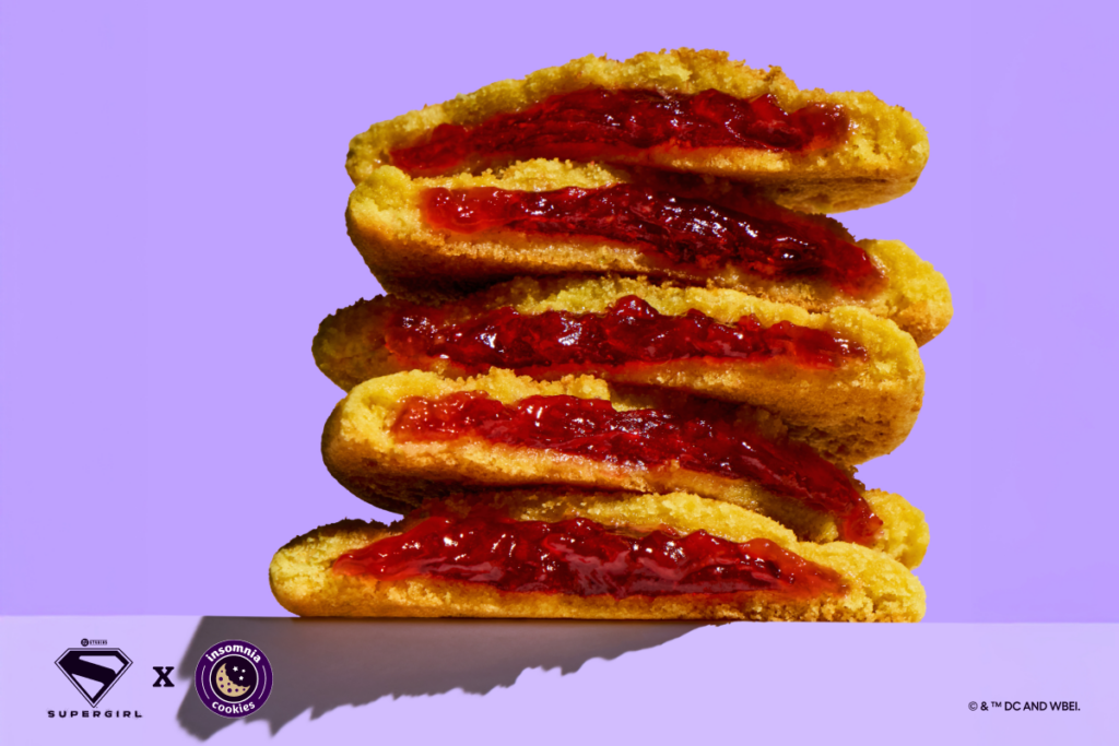 A stack of five halved Lemon Solar Punch cookies filled with bright red strawberry jam, set against a solid pastel purple background. The bottom edge shows a clean white surface featuring a black Supergirl "S" shield logo and the purple Insomnia Cookies logo.