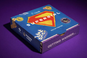 A closed, square Supergirl Insomnia Cookies delivery box sitting on a deep purple background. The top of the box features a blue limited-edition Supergirl sleeve with a large yellow and red Supergirl "S" shield logo in the center, decorated with graphic stickers and the text "ONLY IN THEATERS" for Supergirl Insomnia Cookies promotion