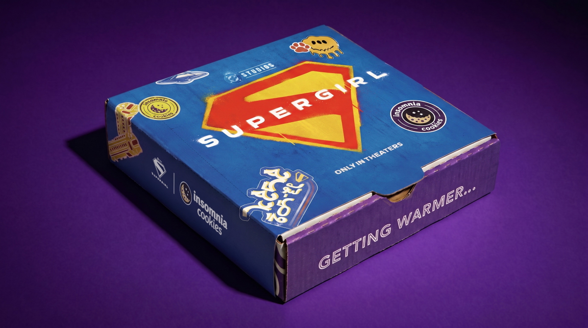 A closed, square Insomnia Cookies delivery box sitting on a deep purple background. The top of the box features a blue limited-edition Supergirl sleeve with a large yellow and red Supergirl "S" shield logo in the center, decorated with graphic stickers and the text "ONLY IN THEATERS" for Supergirl Insomnia Cookies promotion
