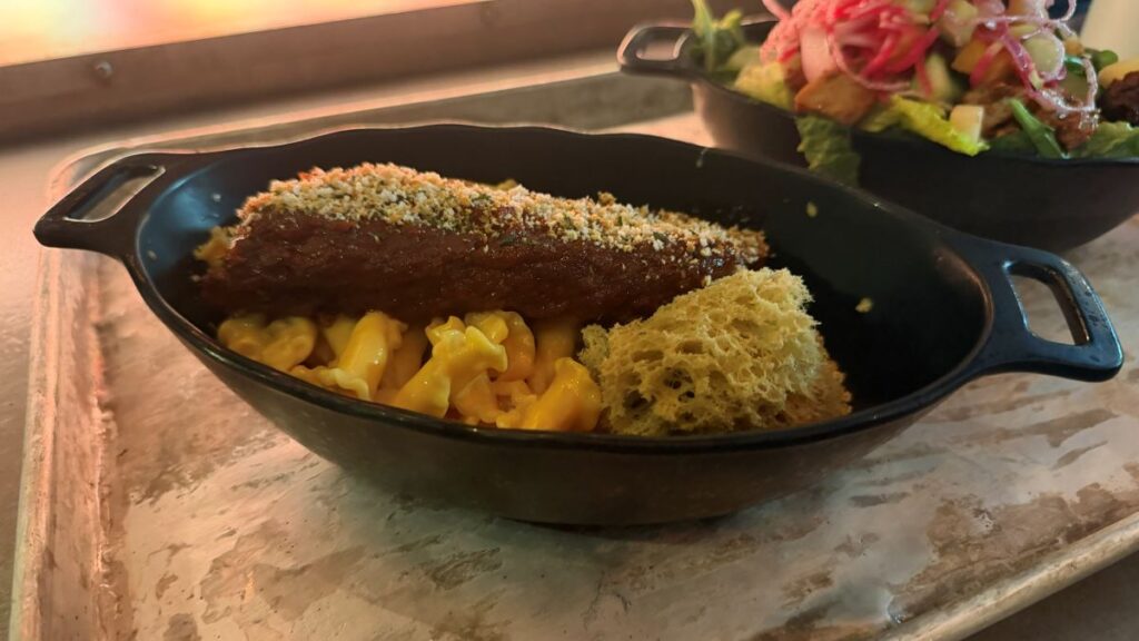 A serving of Sweet Puffer Pig Ribs featuring a large, sauce-covered rib over creamy macaroni and cheese, accompanied by a piece of porous, greenish-yellow portion bread in a black serving dish.