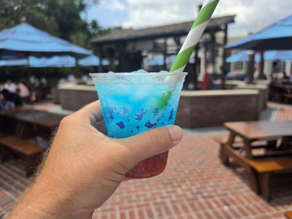 A close-up of a hand holding a bubbly blue and red layered holiday drink with a green-and-white striped straw, perfect as colorful Fourth of July Treats in a brick courtyard setting.